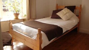 Uma cama de madeira com roupa de cama branca e castanha, uma janela com uma planta e chão de madeira.