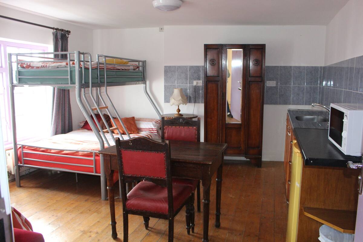 A room with bunk beds, a wooden table with chairs, a lamp, a wooden wardrobe, and a kitchen area with a microwave.