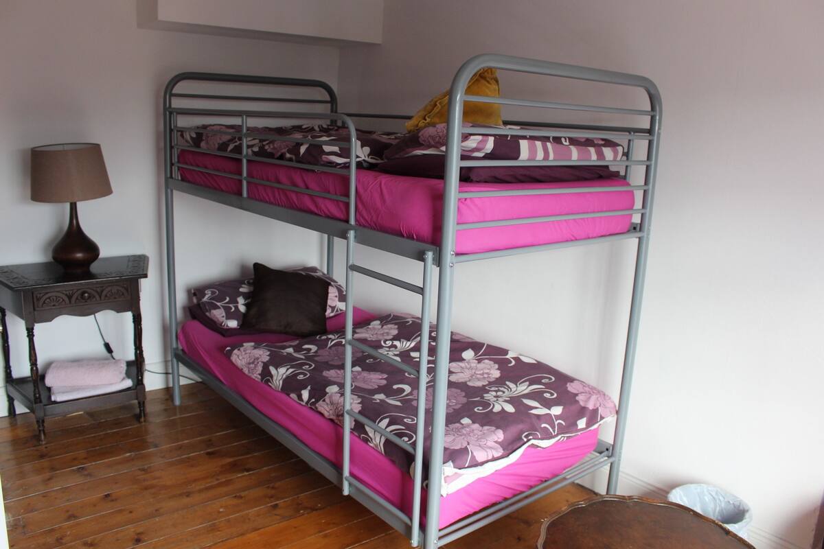 A bunk bed with pink bedding, a small table with a lamp, and a wooden floor.