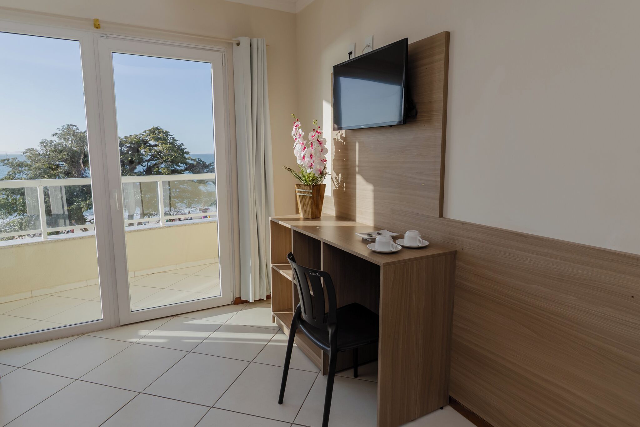 A room with a wooden desk, a chair, a TV mounted on the wall, and a window with a view of trees and the sea.