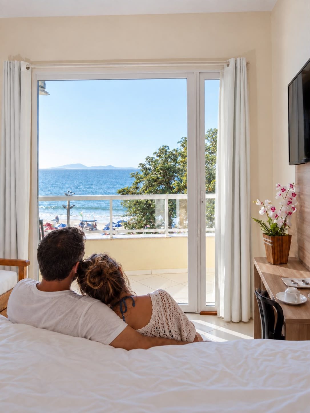 A couple lying on a bed facing a balcony with a view of the sea and beach.
