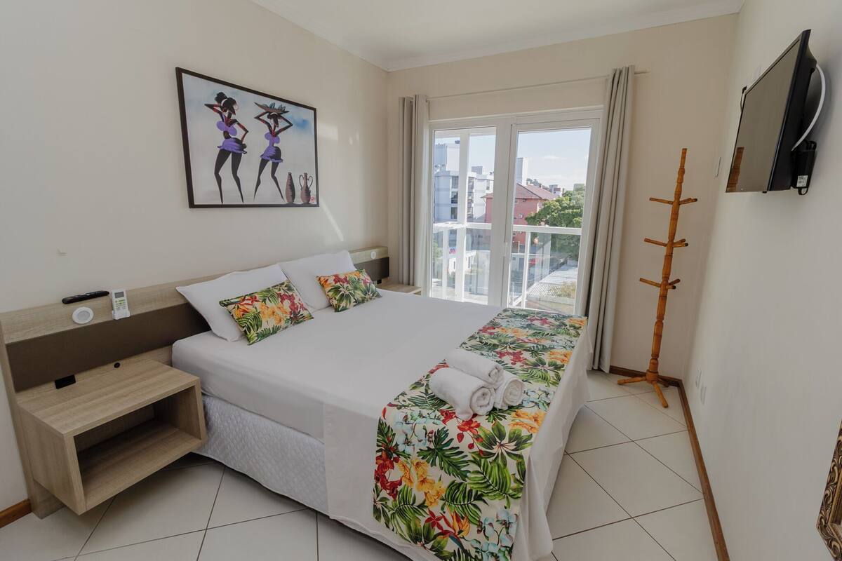 A hotel room with a bed, a bedside table, a TV, and a balcony with a view of buildings.