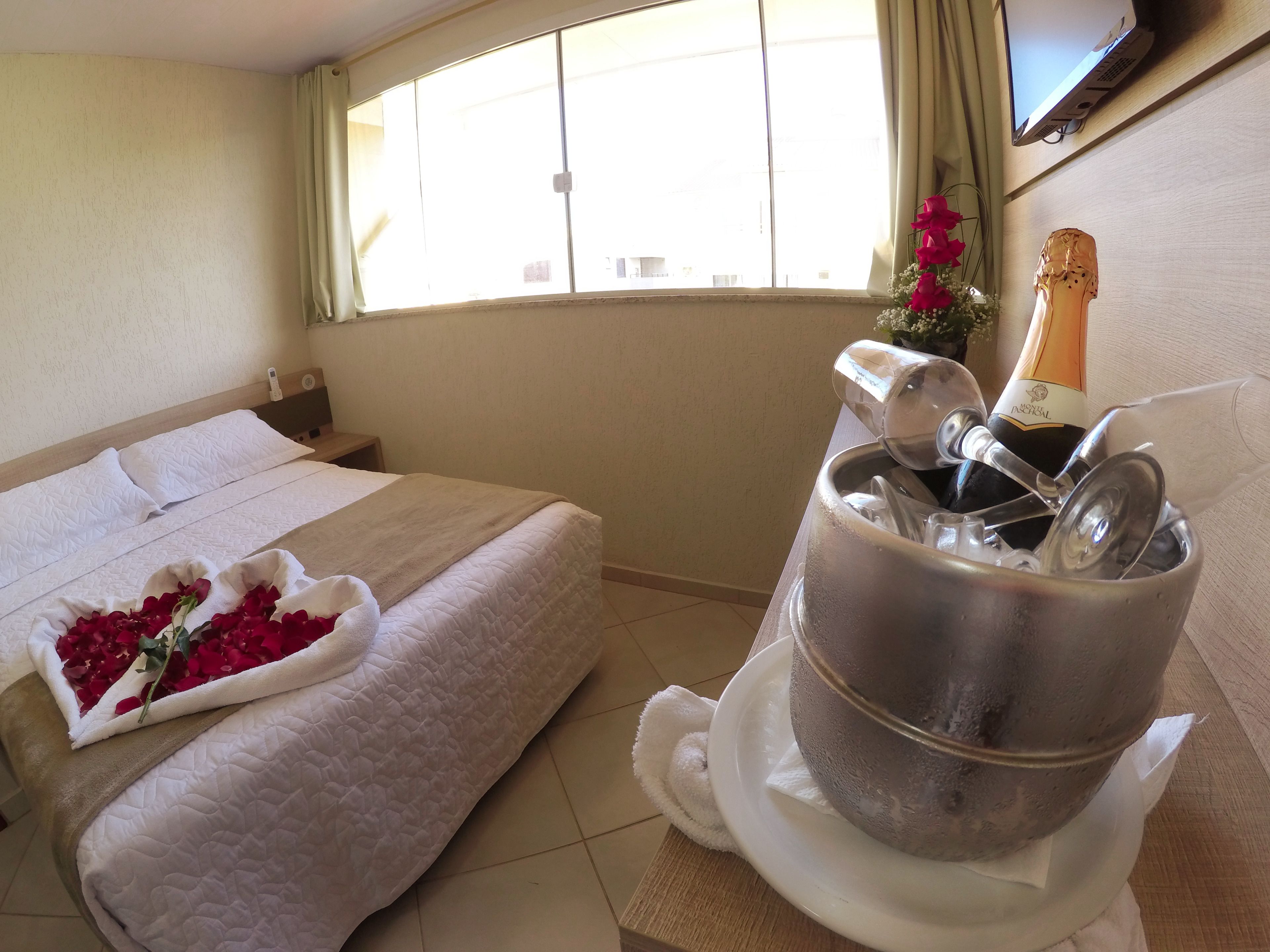 A hotel room with a bed, a bedside table, a window with curtains, a television, and a minibar with a bottle of champagne.