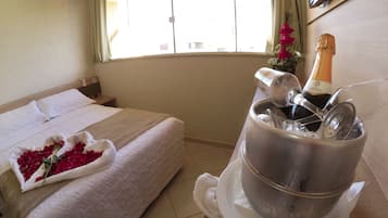 A hotel room with a bed, a bedside table, a window with curtains, a television, and a minibar with a bottle of champagne.