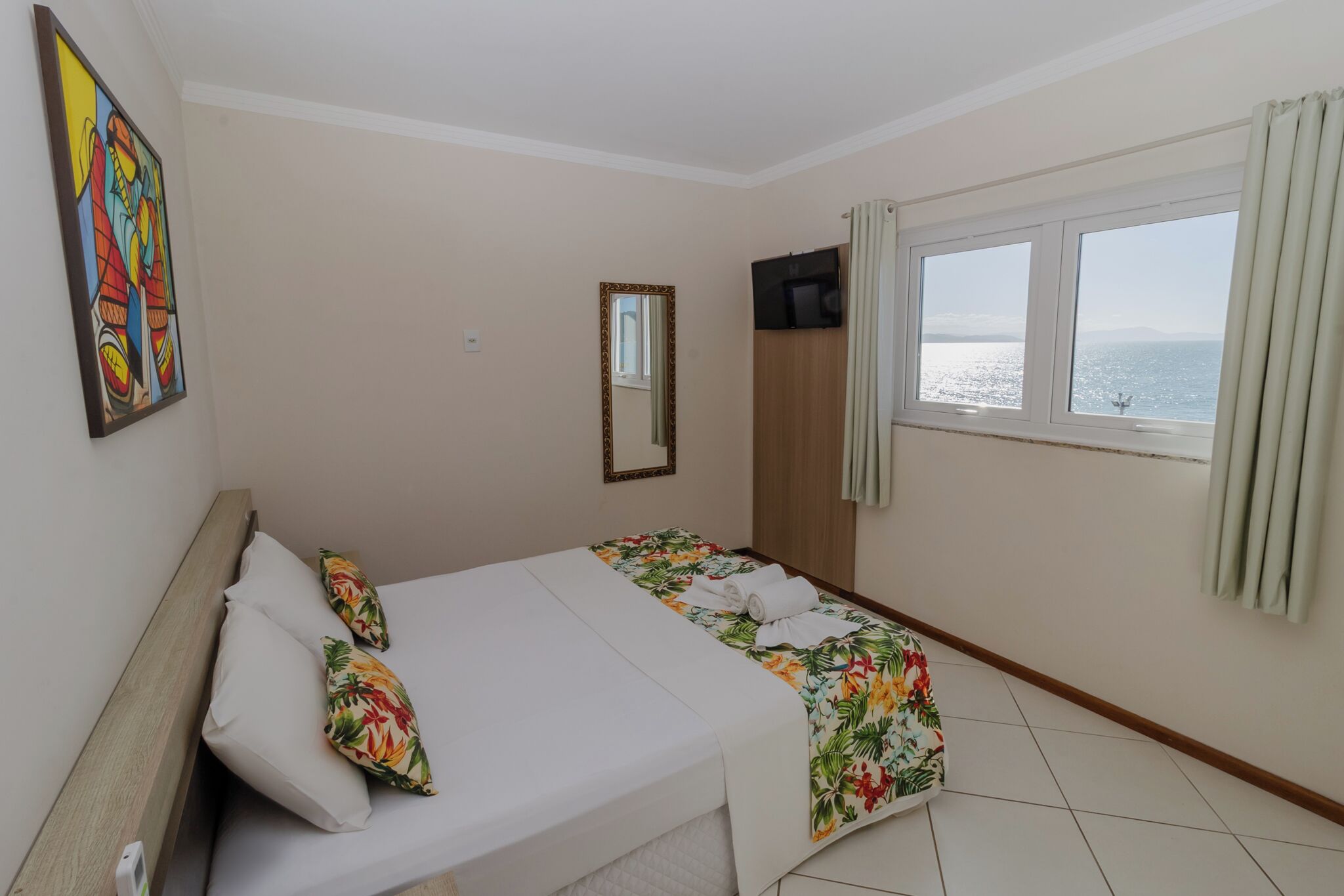 A hotel room with a bed, a window with a view of the sea, a painting on the wall, and a mirror.
