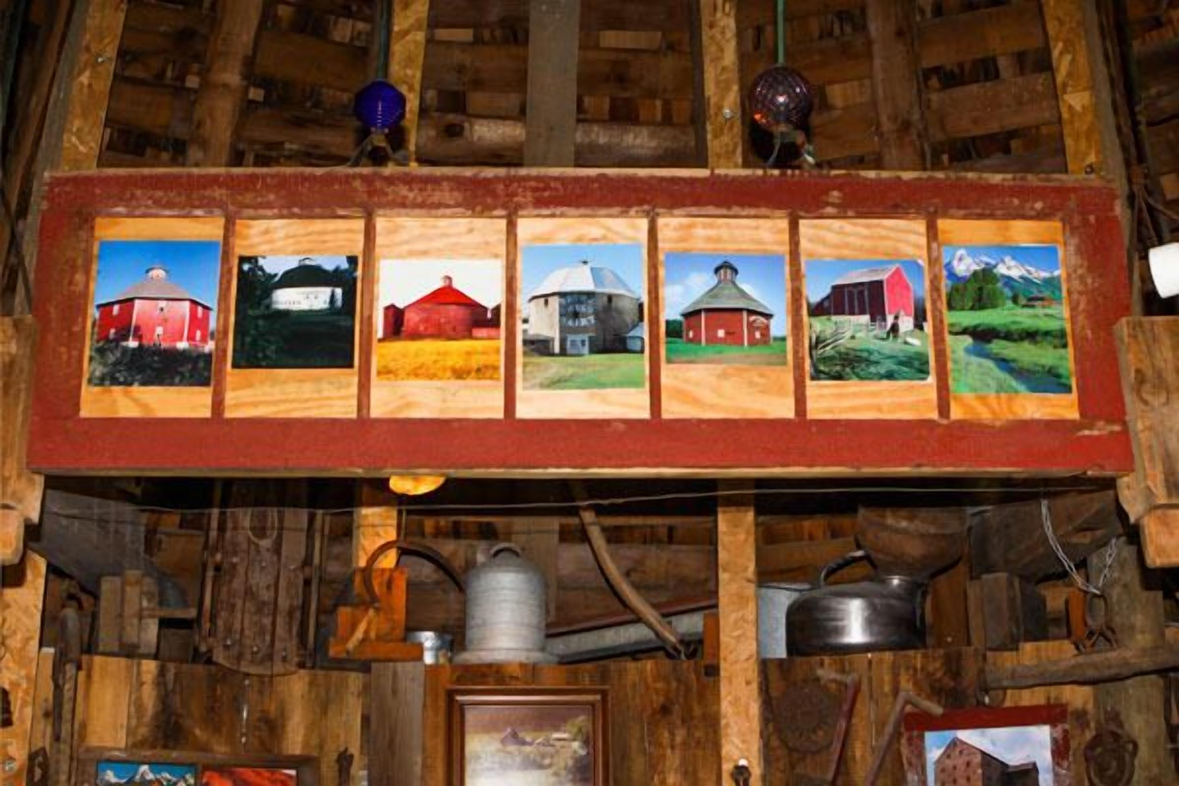 L’intérieur d’une grange rustique, avec des poutres en bois, une étagère en bois où sont exposées six photographies encadrées de granges, ainsi que divers objets agricoles, comme une citerne à eau et des pots.