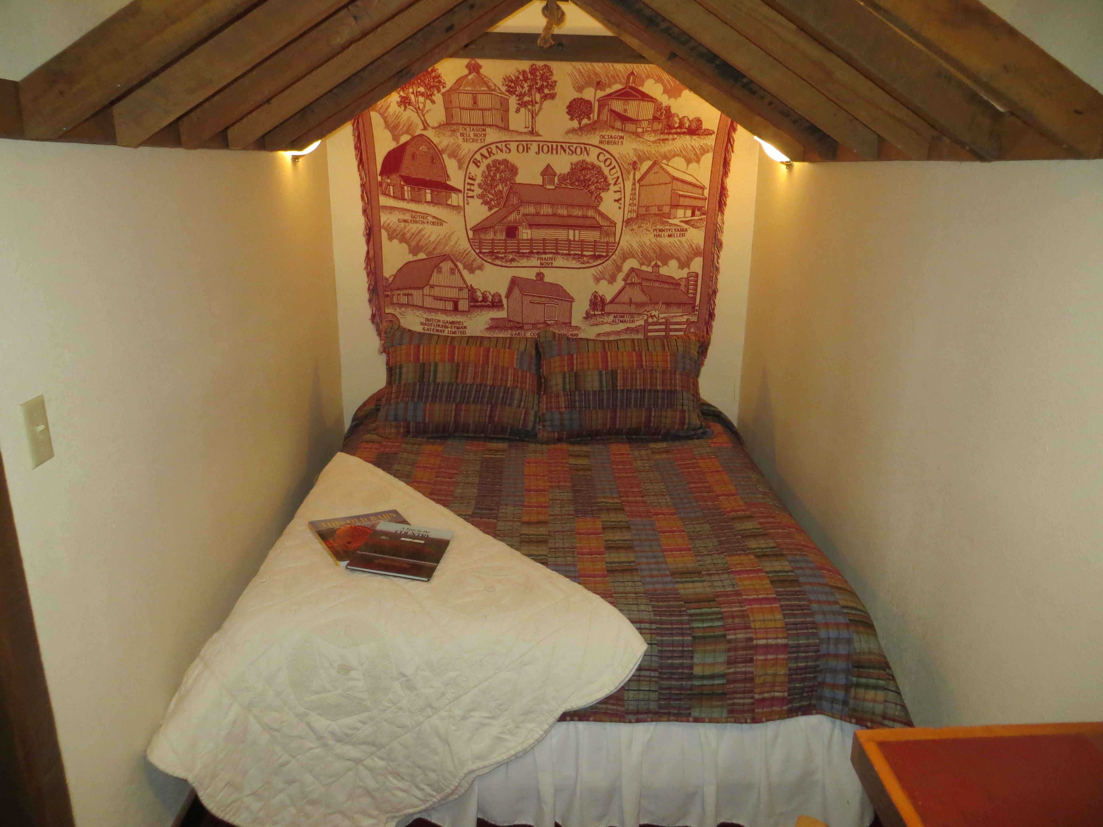 A small, cozy room with a bed, a quilted blanket, and a tapestry depicting local barns and landscapes.