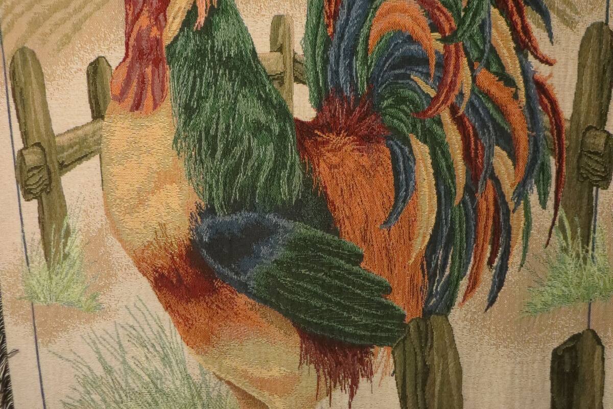 A rooster standing on a fence with a rural landscape in the background.