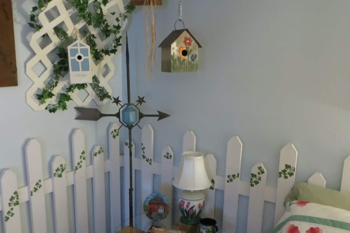 A room with a white picket fence, a wooden table with a lamp, a book, and a decorative birdhouse.