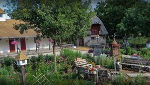 A rustic village scene with traditional buildings, a thatched-roof house, and a garden with various plants and flowers. - Cupola Inn Bed & Breakfast (Nora Springs)