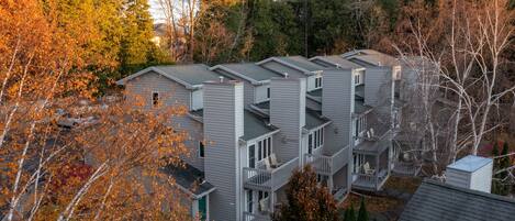 A row of modern, multi-story apartment buildings with balconies, surrounded by autumn foliage.