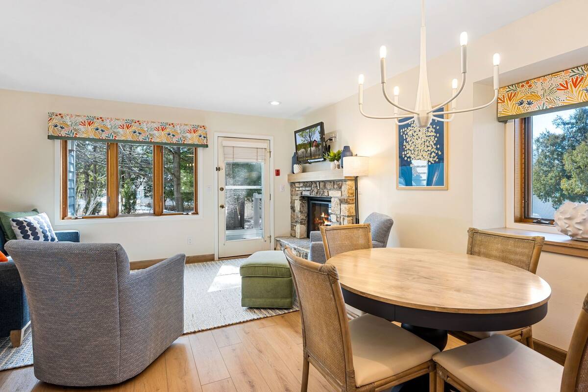 A dining area with a round table and chairs, a fireplace, and a view of trees through the windows.