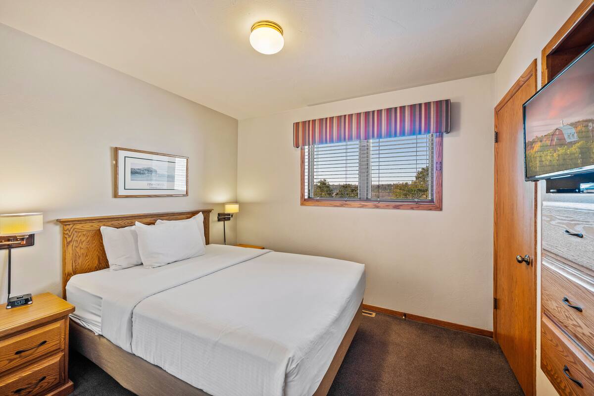 A neatly made bed in a hotel room with a wooden headboard, a bedside table with a lamp, a framed picture on the wall, and a window with blinds.
