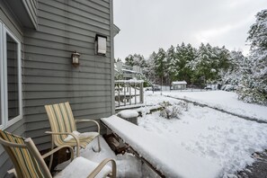 A covered outdoor seating area with chairs and a table, adjacent to a house with a window and a wall-mounted light fixture. - Evergreen Hill Condominiums (Fish Creek)