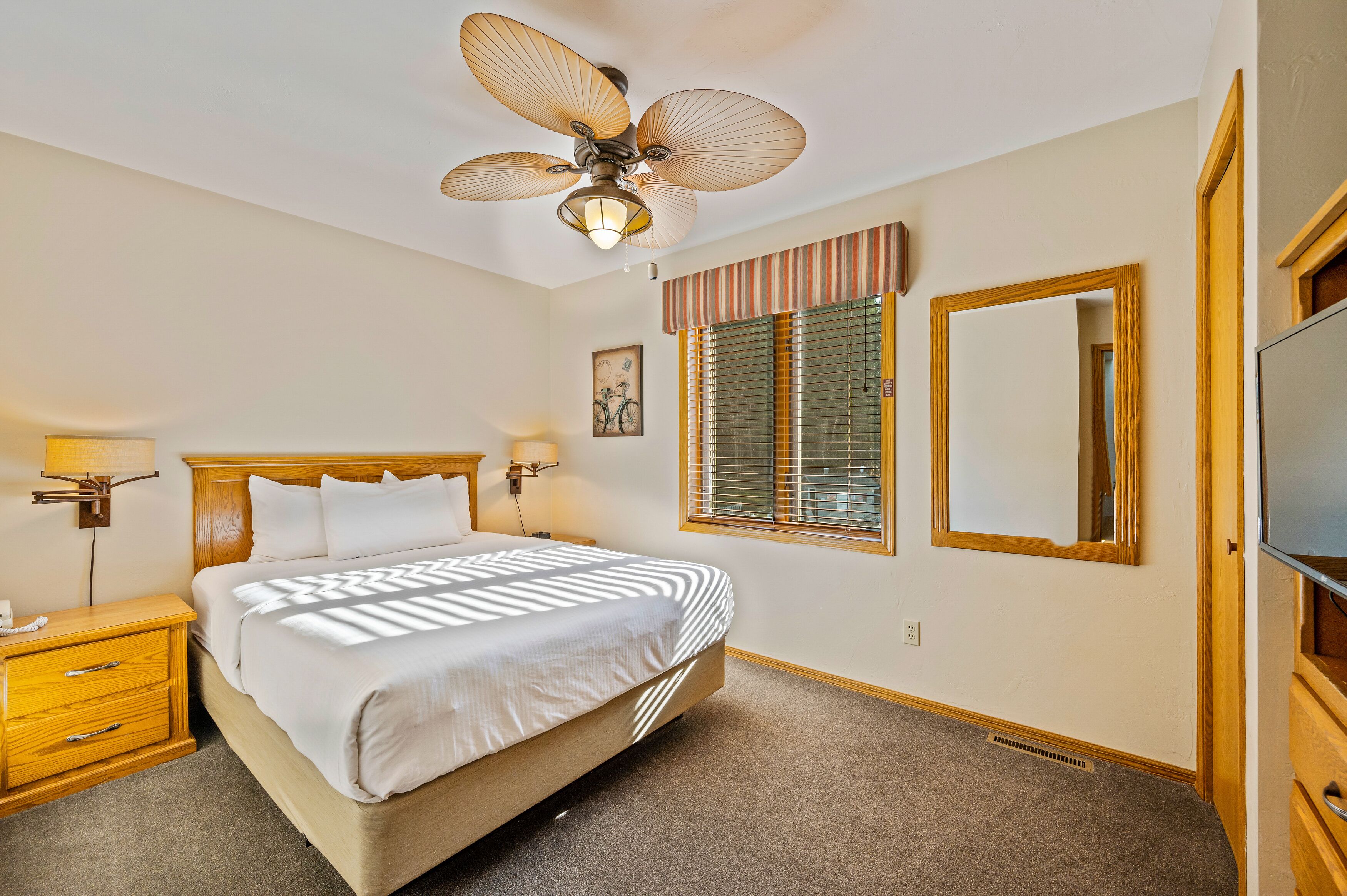 A neatly arranged bedroom with a bed, bedside tables, a ceiling fan, a window with blinds, a mirror, and a television.