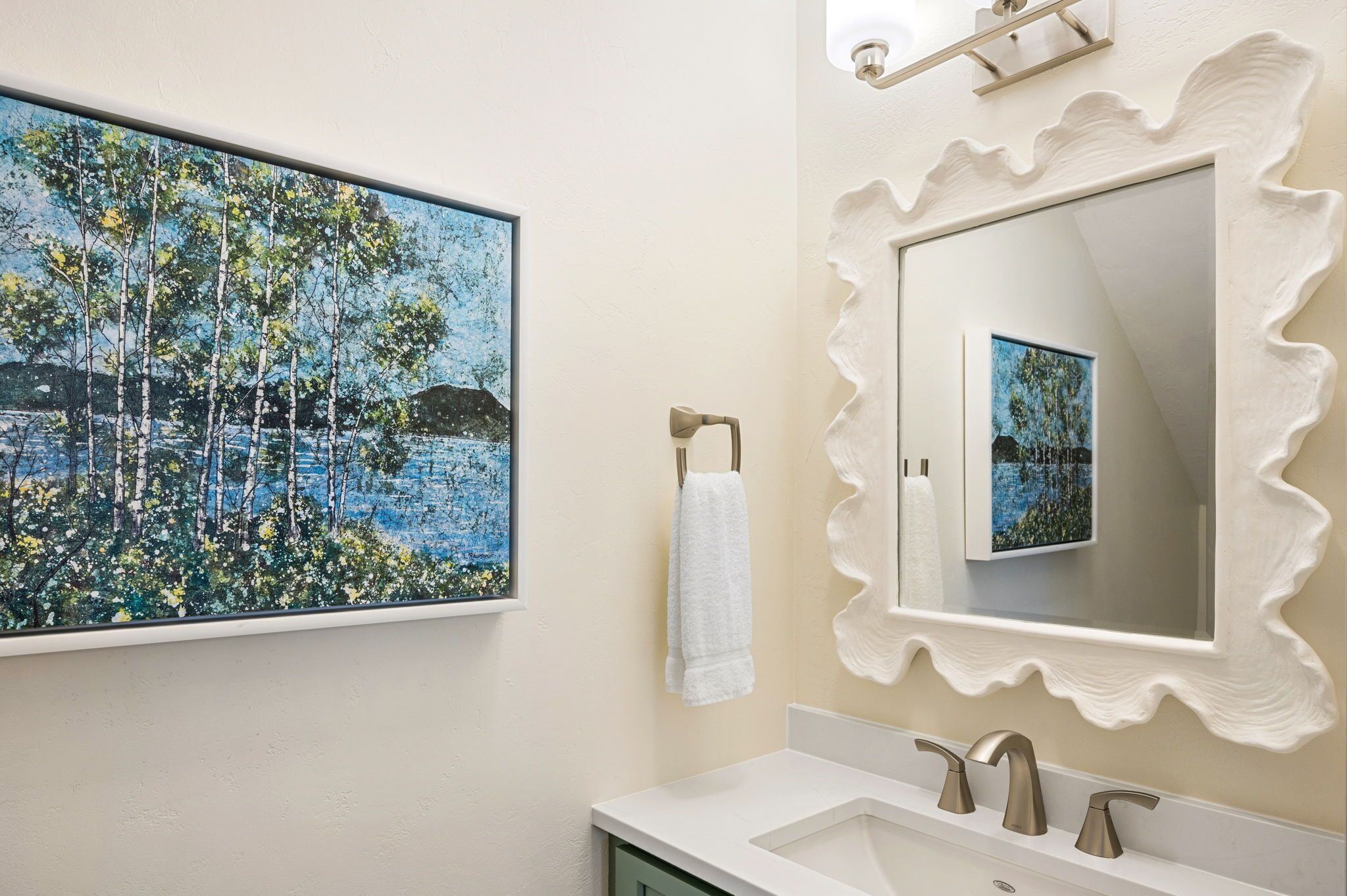 A bathroom with a sink, two faucets, a white towel, a painting of a forest scene, and a mirror with a wavy frame.