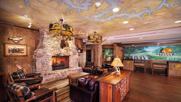 Interior rústico de una cabaña con chimenea de piedra, muebles de madera, adornos de asta de ciervo y una gran pantalla que muestra un campo de golf.