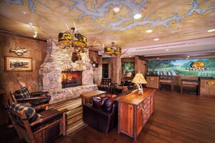 A rustic lodge interior with a stone fireplace, wooden furniture, antler decorations, and a large screen displaying a golf course.