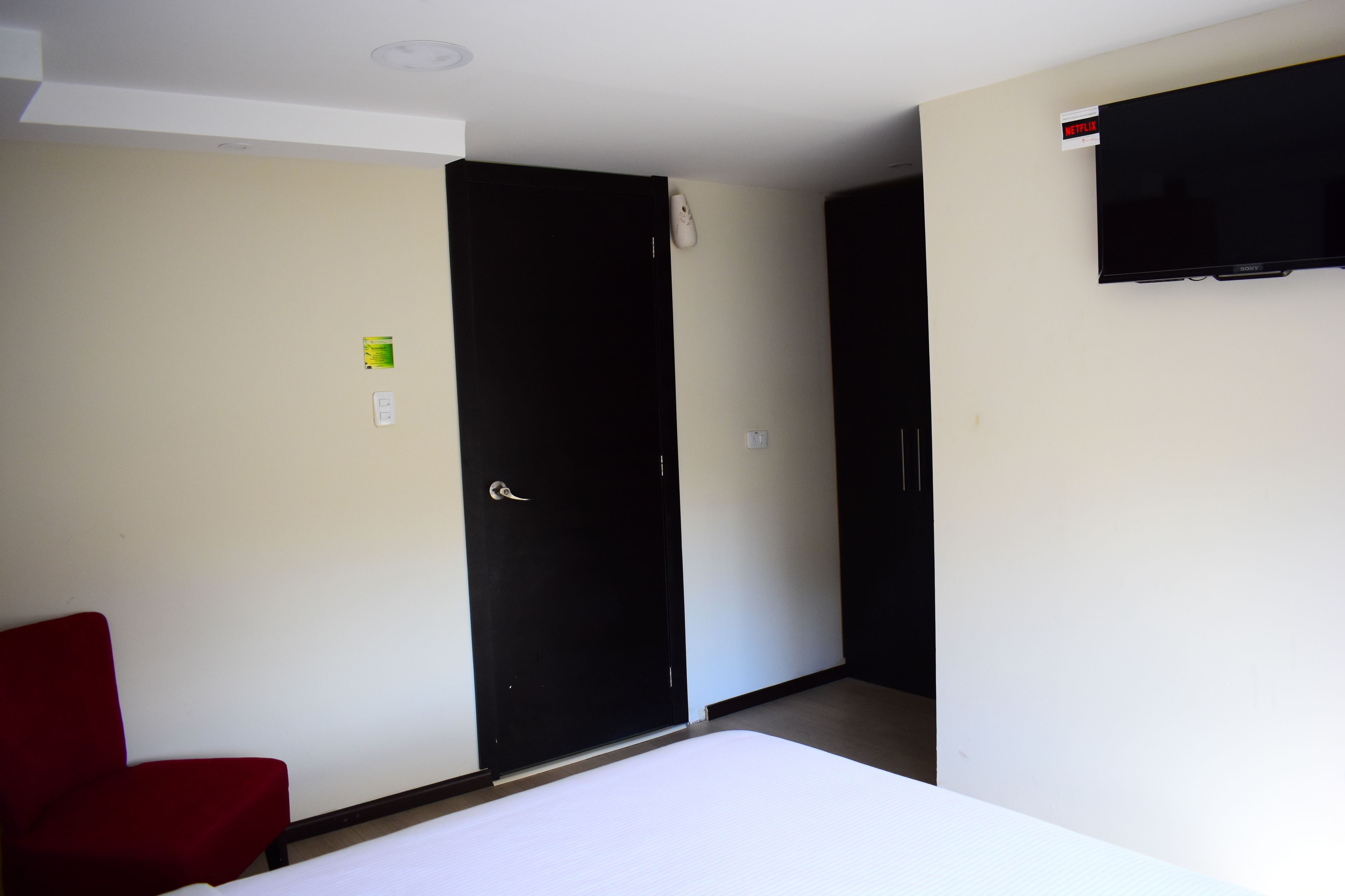 Et hotelværelse med en seng, en rød stol, en sort garderobe og et fjernsyn på væggen.