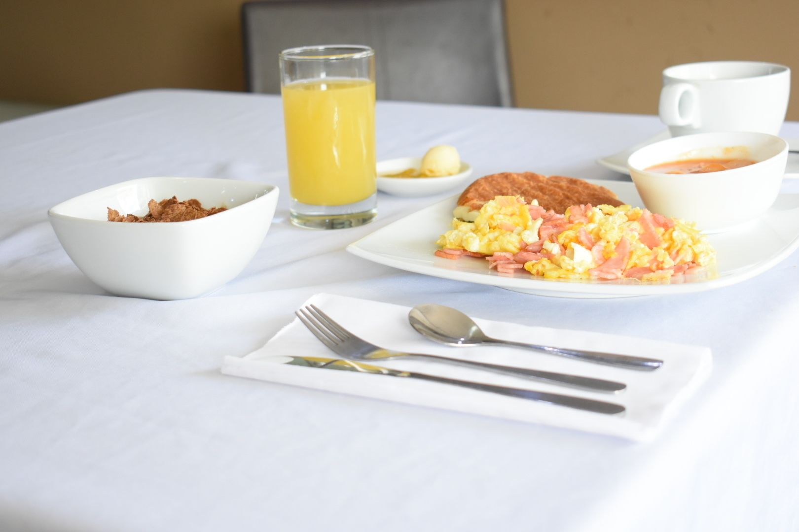 Una mesa de desayuno con huevos revueltos, tostadas, un vaso de jugo de naranja, un tazón de cereal y una taza de café.