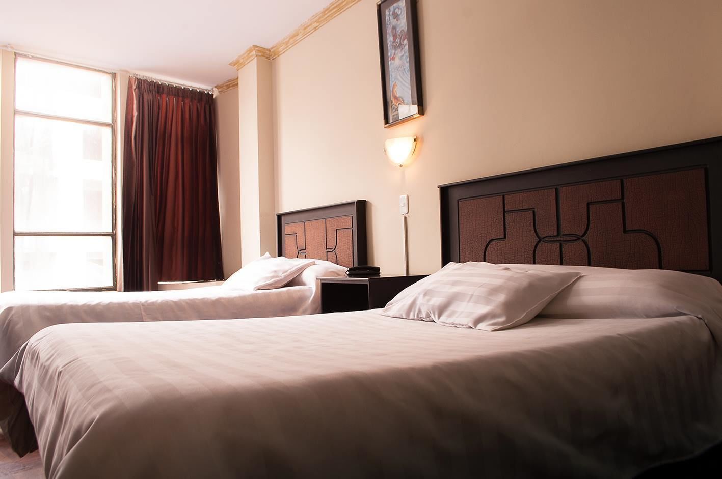 A hotel room with two beds, a nightstand, a lamp, and a window with curtains.