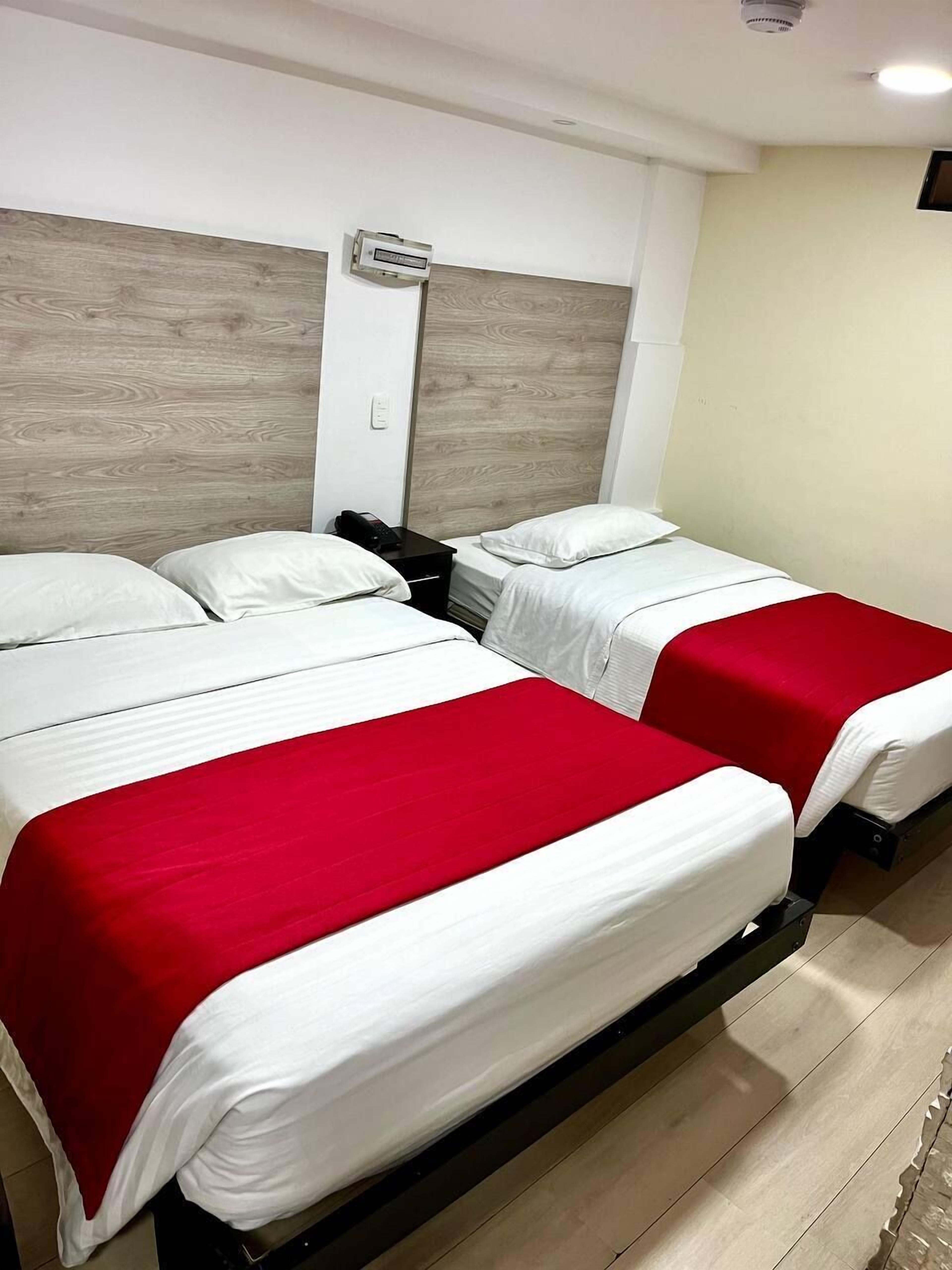 A hotel room with two beds, each with white bedding and red accents, a wooden headboard, a bedside table with a phone, and a wall-mounted air conditioning unit.