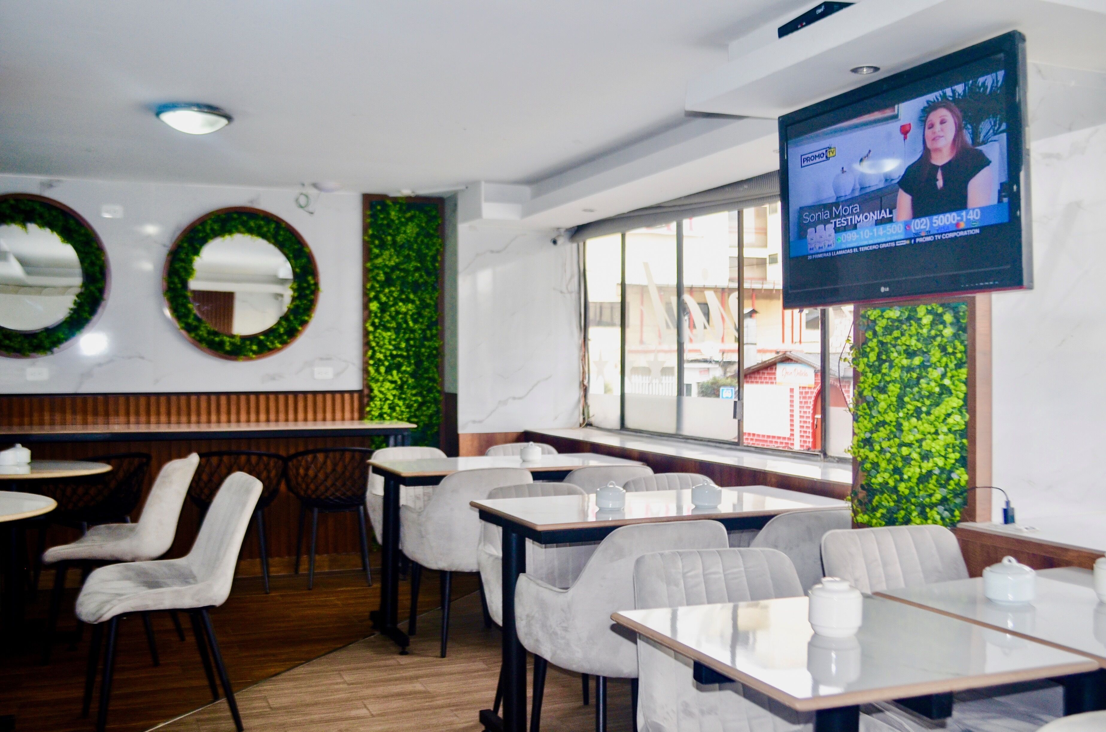 Interior de restaurante com mesas redondas, cadeiras, uma tela de TV exibindo um programa e decorações verdes nas paredes.