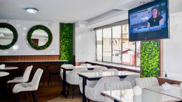 Interior de restaurante com mesas redondas, cadeiras, uma tela de TV exibindo um programa e decorações verdes nas paredes.