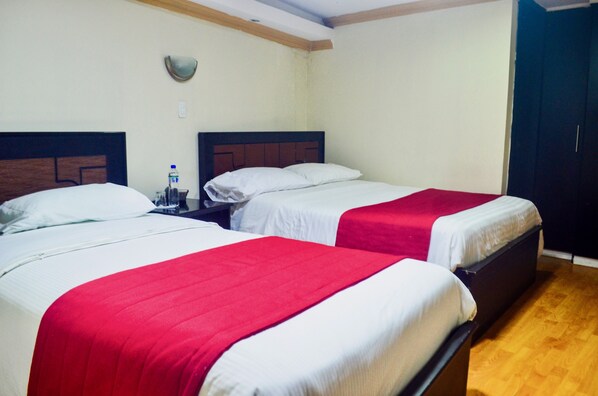 A hotel room with two beds, each with a red and white bedspread, a nightstand with a bottle, and a wooden floor.