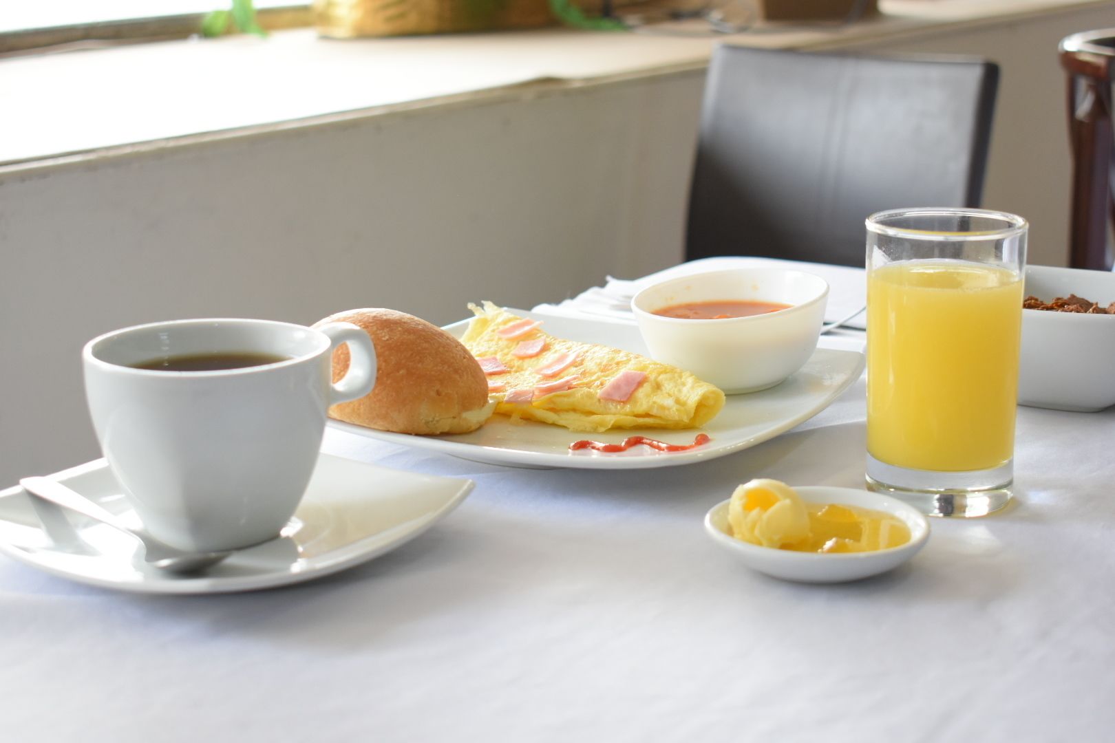Desayuno con una taza de café, un croissant, un tazón de sopa, un vaso de jugo de naranja y un recipiente pequeño de mantequilla sobre una mesa.