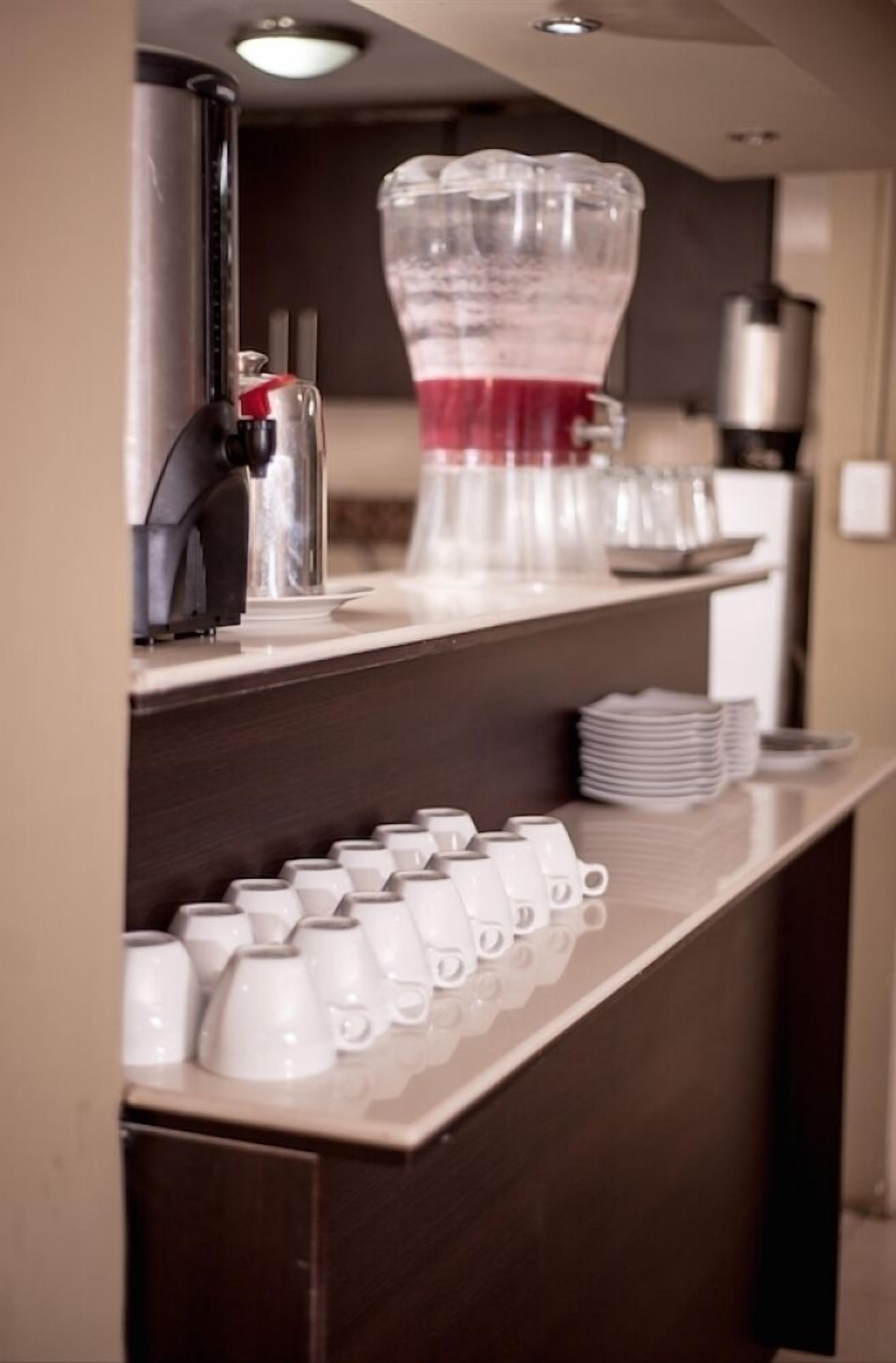 Una zona de bar con una máquina de café, un recipiente de vidrio grande con líquido rojo y varias tazas y vasos blancos dispuestos en estantes.