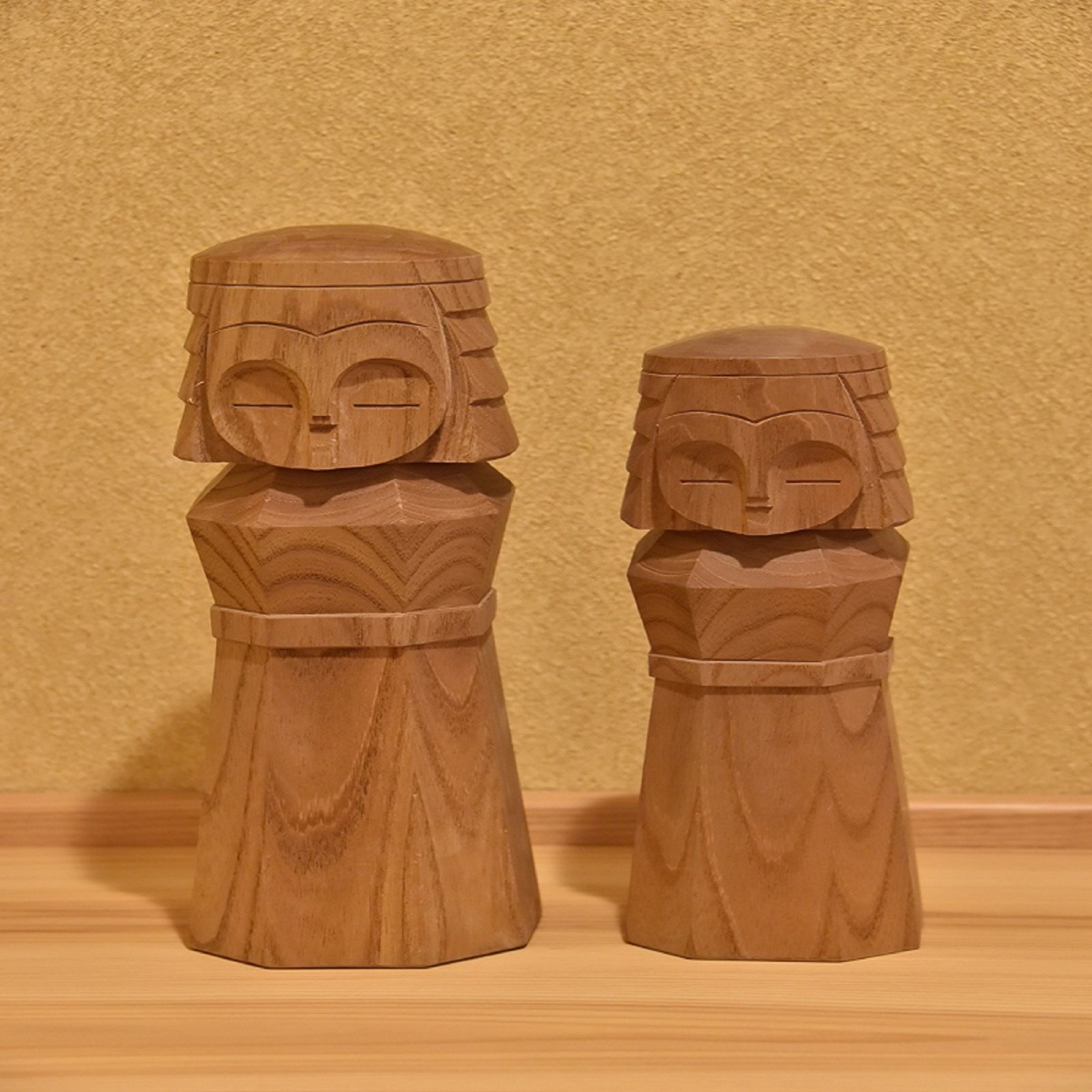Two wooden sculptures with faces on their heads, placed on a wooden surface against a plain wall.