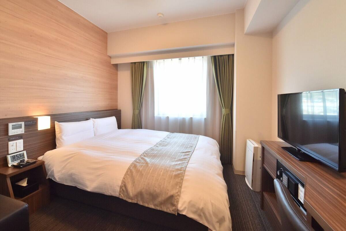 Una habitación de hotel con cama, mesitas de noche, televisión y ventana con cortinas.
