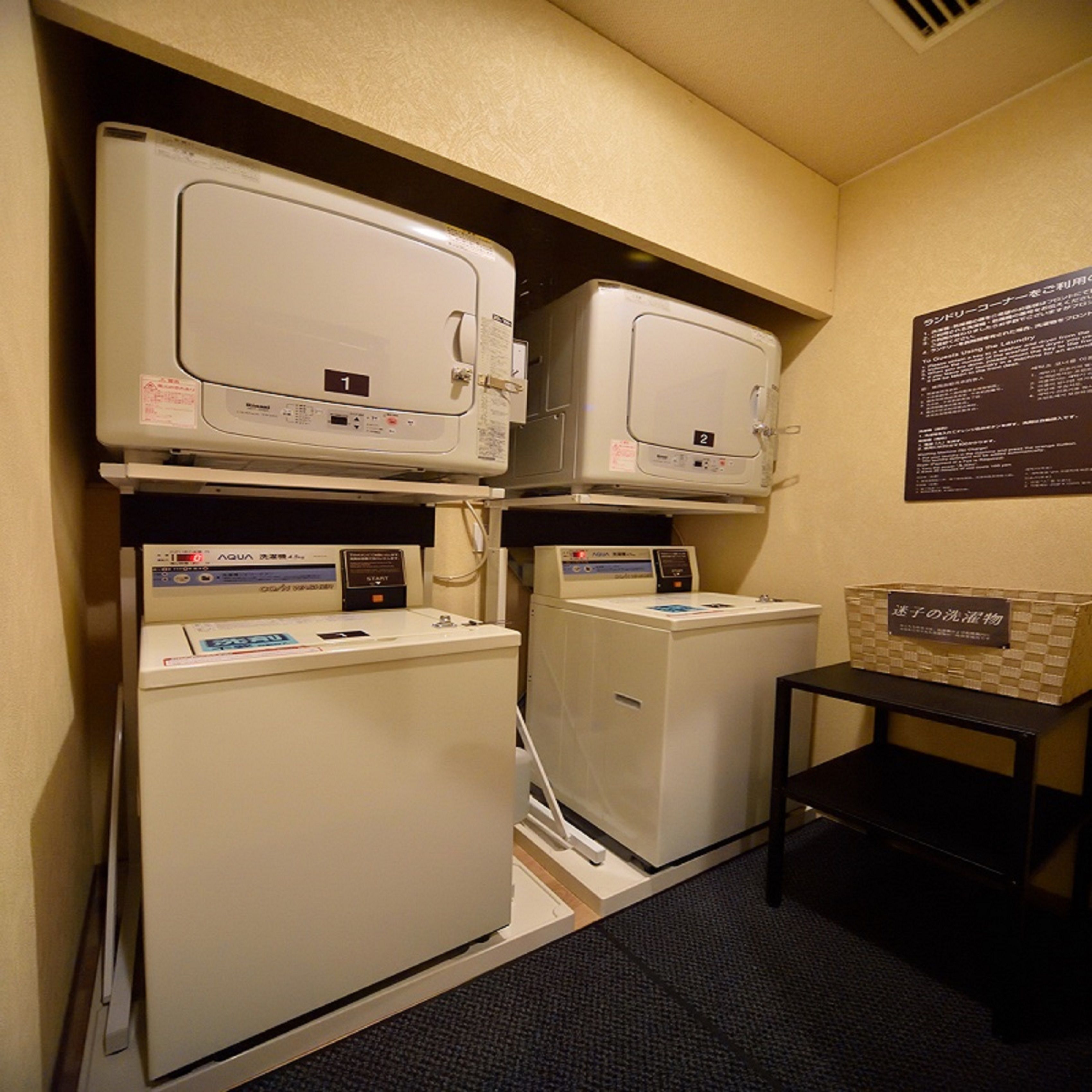 A laundry facility with two dryers and a washing machine, a small table with a basket, and a sign with instructions.