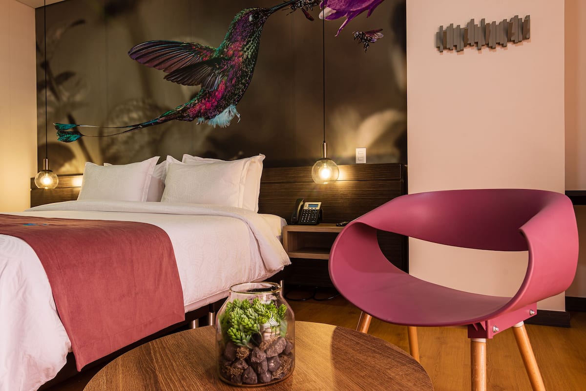 A modern hotel room with a bed, a glass jar with succulents, a pink chair, and a decorative hummingbird.