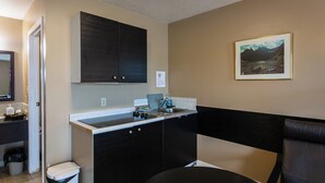 A compact kitchen with dark cabinets, a sink, and a countertop. There is a trash can and a chair visible. - Chinook Country Inn (Sundre)