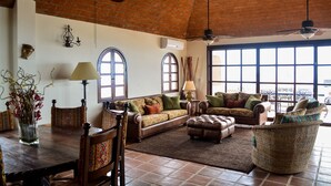 A spacious living room with a high ceiling, large windows, and a dining area with a table and chairs. - Hacienda Edith (Cabo San Lucas)