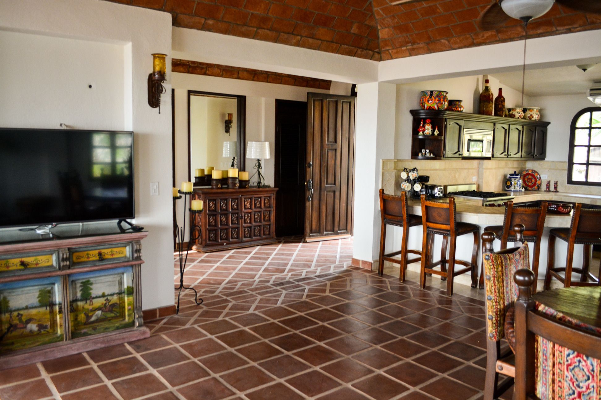 Amplia sala de estar con suelo de baldosas, televisor, mueble de madera y una zona de cocina con taburetes de bar.