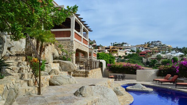 A stone staircase leading to a pool area with lounge chairs and a view of a hillside residential area. - Hacienda Edith (Cabo San Lucas)