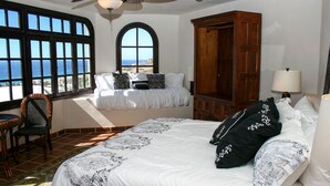 A bedroom with a large bed, a wooden wardrobe, a chair, and a view of the ocean through large windows. - Hacienda Edith (Cabo San Lucas)