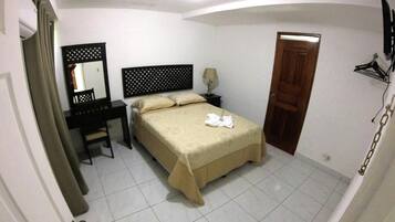 A bedroom with a bed, a desk, a chair, a mirror, a nightstand, a lamp, and a door.