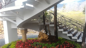 Uma escadaria em espiral com corrimão de metal, cercada por um jardim com flores vermelhas e arbustos verdes.
