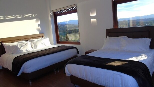 Duas camas de solteiro com cabeceiras de madeira e roupa de cama branca, uma janela grande com vista para montanhas e um painel decorativo de madeira.