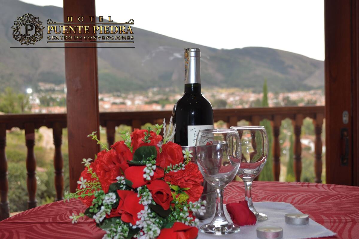 Une table dressée avec une bouteille de vin, deux verres à vin et un bouquet de roses rouges, sur un balcon donnant sur une ville et des montagnes.