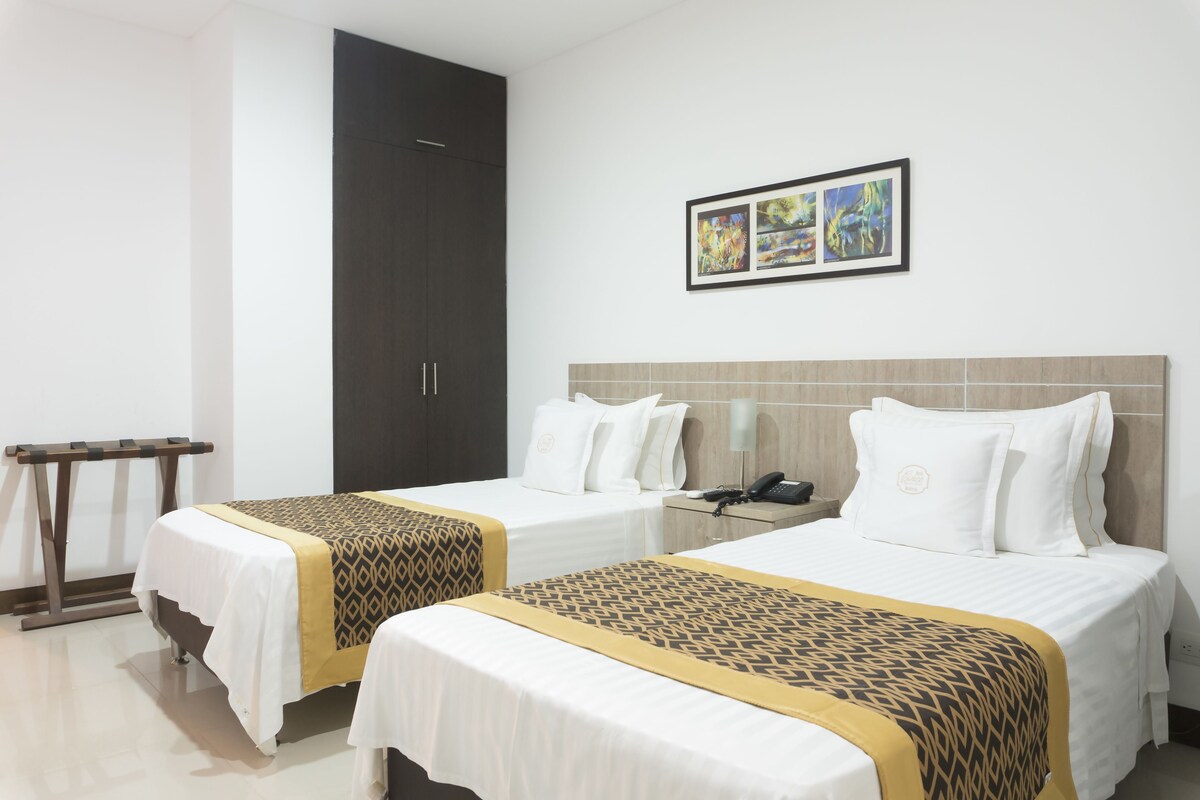 Hotellihuone, jossa on kaksi sänkyä, vaatekaappi, yöpöytä lampun kanssa ja taideteoksia seinällä.