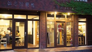 A brick building with large glass doors and windows, a sign reading "Guest House," and outdoor seating with tables and chairs. - BACCHUS Kofu Guest House - Hostel (Kofu)