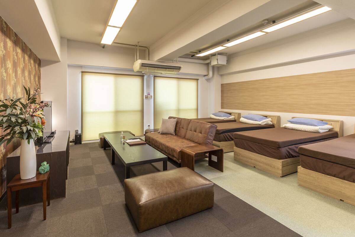 A modern hotel room with a sofa, two single beds, a wooden headboard, a glass coffee table, a small table with a laptop, and a potted plant.