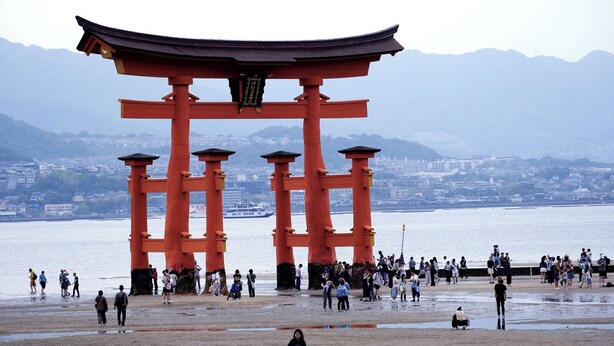 A traditional Japanese torii gate with a cityscape and mountains in the background. - OMOTENASHI Hostel Miyajima (Hatsukaichi)