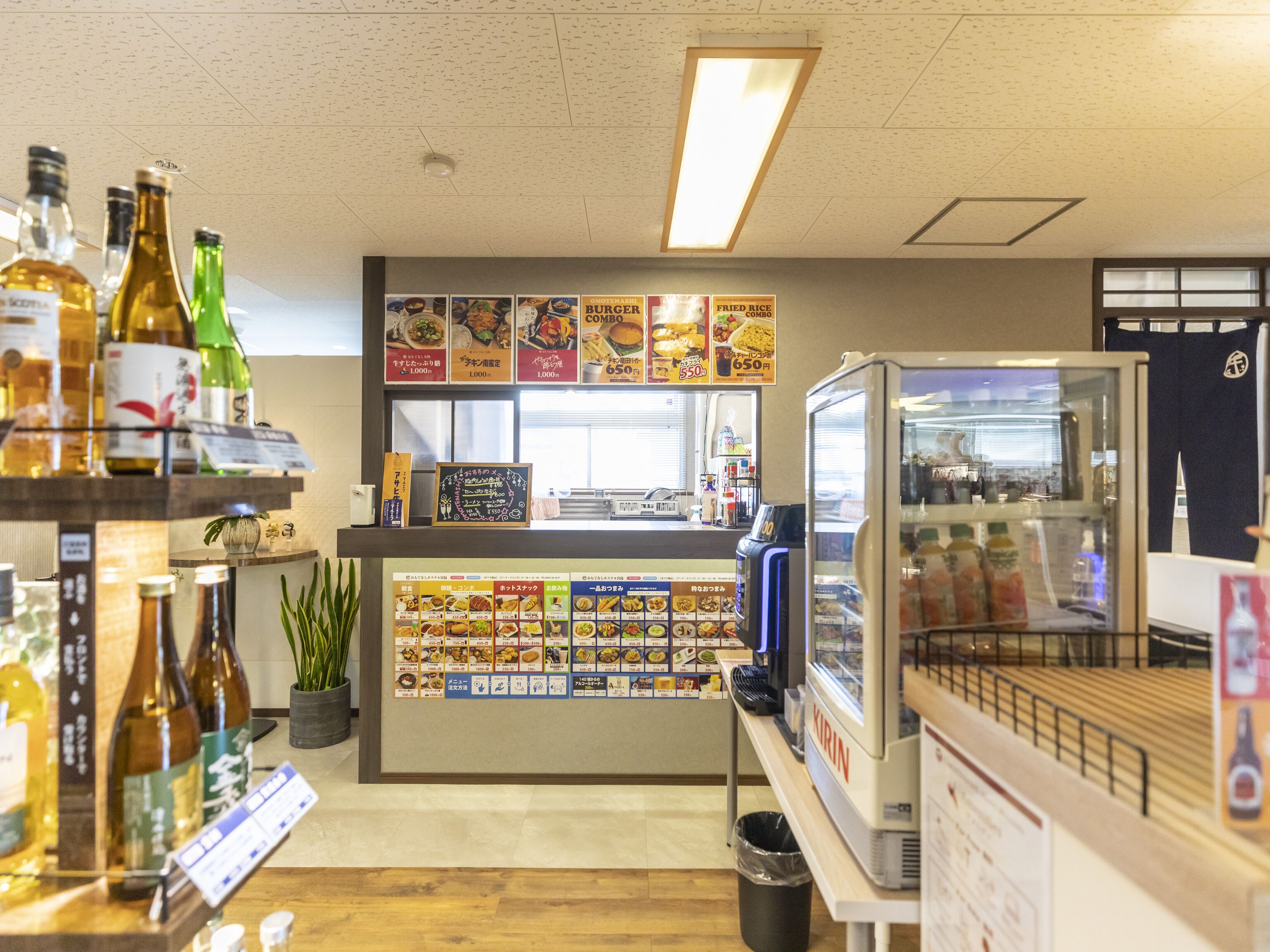 Interior de restaurante con mostrador que exhibe alimentos, vitrina refrigerada y estantes con botellas.