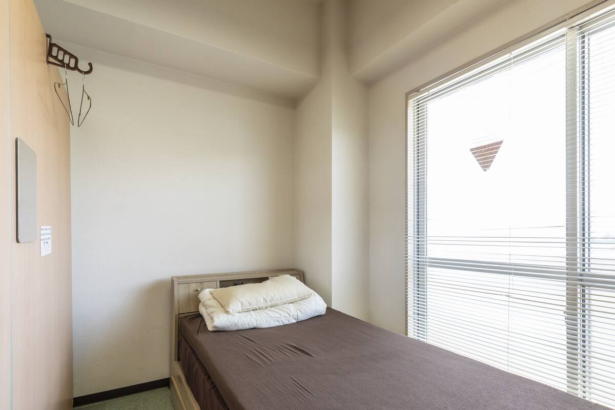 A small, single-bed room with a wooden headboard, a brown mattress, and a white pillow. There is a window with blinds on the right side, and a hook on the wall to the left.